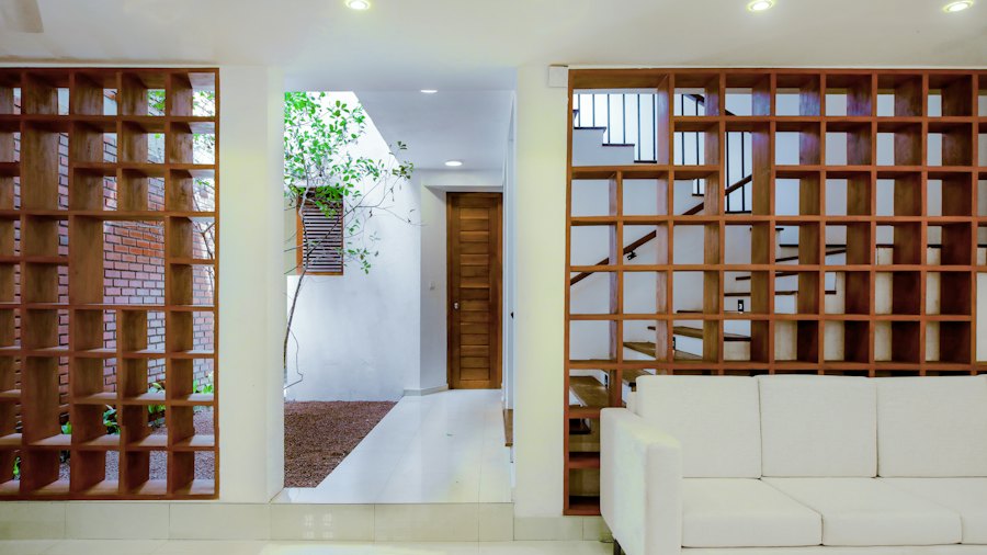 Sala com sofá branco entre duas estantes de madeira em treliça; ao fundo, a escada e a luz que vem do jardim interno.