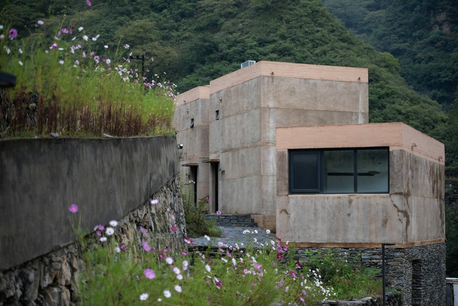 Volumes de concreto em tom de terra apoiados na encosta, com muro de pedra e jardim de flores em primeiro plano.